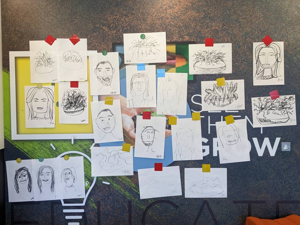 Wall covered in charcoal sketches on pieces of paper. They vary in style and are a mix of faces and plants.