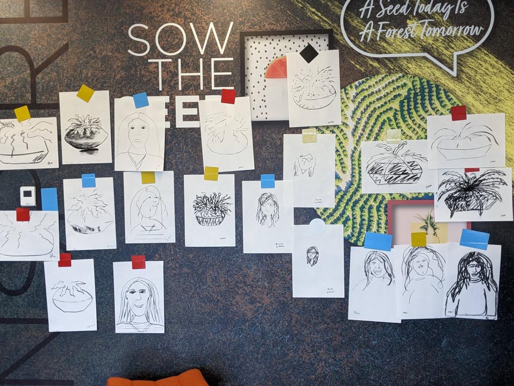 Wall covered in charcoal sketches on pieces of paper. They vary in style and are a mix of faces and plants.